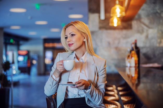 Blonde professionnelle d'âge moyen assis dans le café d'un hôtel et tenant une tasse de café blanc. Elle attend quelqu'un. Plaisir, pause café