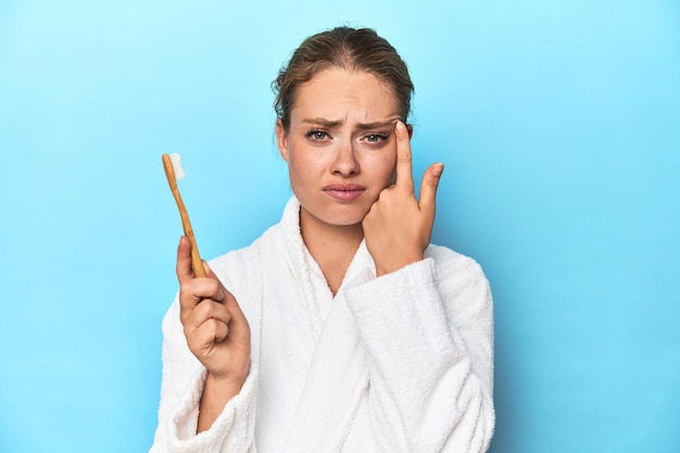 Blonde en robe de bain avec une brosse à dents dans un studio bleu montrant un geste de déception avec l'index