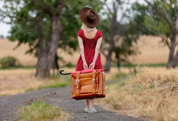 Blonde en robe rouge avec une valise sur une route rurale avant la pluie