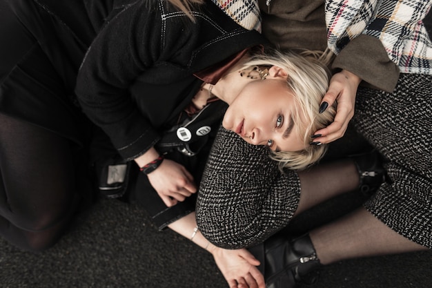 Blonde sexy de jeune femme dans des vêtements élégants noirs reposant sur l'asphalte avec la soeur dans la veste à carreaux sur la rue