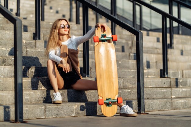 Blonde sexy avec un longboard est assise dans les escaliers en short court. Photo de haute qualité