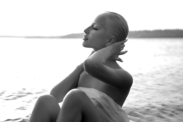 Une blonde sexy nue avec une coupe de cheveux courte est assise dans l'eau sur la plage du lac au coucher du soleil. Cheveux mouillés et corps de femme. Vacances à la plage isolée. Noir et blanc