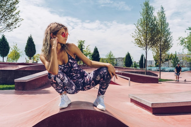 Blonde sportive dynamique en lunettes de soleil dans une pose de prédateur accroupie sur une petite forme architecturale dans le parc