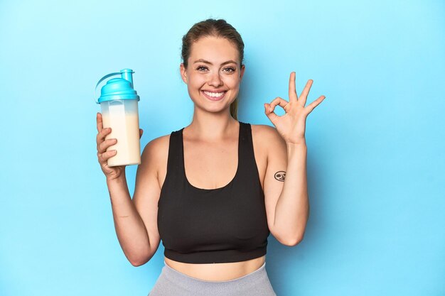 Une blonde sportive avec un shake protéique dans un studio joyeuse et confiante montrant un geste ok