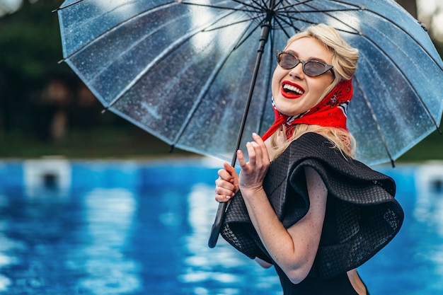 Blonde de style avec des lunettes de soleil tenant un parapluie dans la piscine