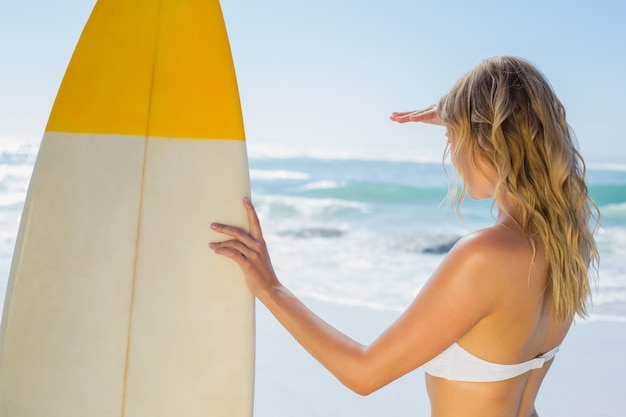 Blonde surfeuse en bikini blanc tenant son tableau sur la plage