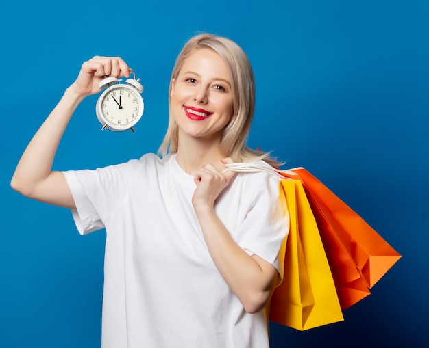 Blonde en t-shirt blanc avec sacs à provisions et réveil sur cintre sur espace bleu