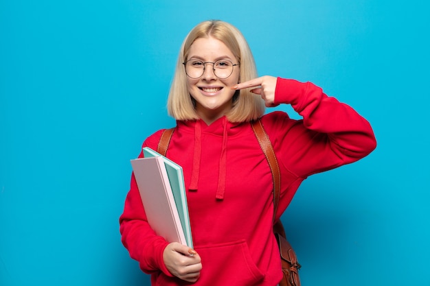 Blonde woman smiling pointant vers son propre large sourire, attitude positive, détendue et satisfaite