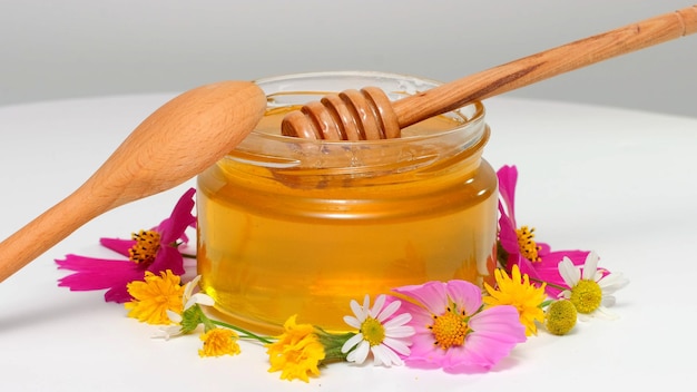 Le bocal en verre avec du miel frais sur fond blanc. Fleurs sur la table. Miel dégoulinant de miel louche dans un bocal en verre. Concept d'alimentation saine, régime alimentaire. Fond de miel