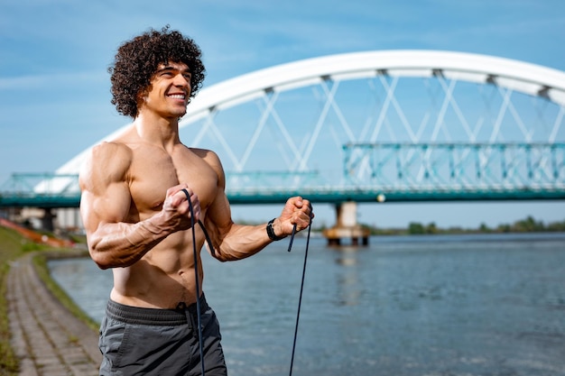 Bodybuilder au torse nu, fait un entraînement corporel solide avec un élastique en caoutchouc, près de la rivière.