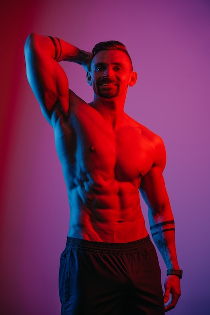 Un bodybuilder avec une barbe pose avec un bras musclé derrière sa tête sous la lumière bleue et rouge