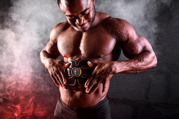 Bodybuilder mâle africain utilise ceinture électronique stimulateur musculaire entraîneur muscles abdominaux sur fond noir avec de la fumée