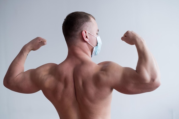 Bodybuilder masculin dans un masque médical se tient avec son dos Le gars fait du sport en quarantaine Protection respiratoire Montre les biceps