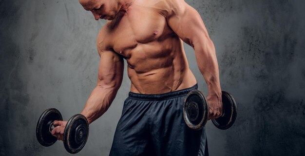 Bodybuilder musclé attrayant fait des exercices avec des barbillons et montre ses muscles sur le fond sombre.