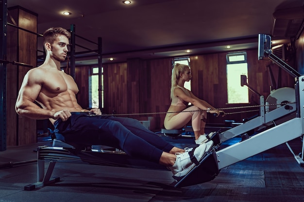 Bodybuilder musculaire dans la formation de vêtements de sport dos assis sur une machine d'exercice. Vue latérale de l'homme avec un corps de musculation parfait dans une salle de sport dans une atmosphère sombre. Concept de musculation, fitness.