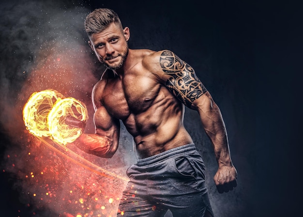 Bodybuilder puissant et élégant avec tatouage sur son bras, faisant les exercices sur un biceps avec des haltères, regardant une caméra. Concept d'art du feu.