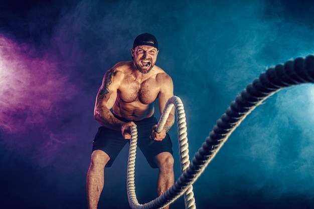 Photo bodybulder à la recherche athlétique barbu s'entraîner avec une corde de combat sur l'obscurité avec de la fumée. force et motivation.