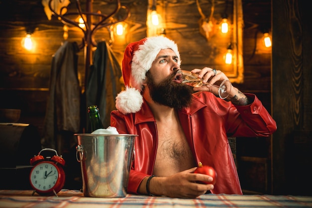 Boire de l'alcool. Bonne année. Veste en cuir de père Noël brutale virile. Père Noël brutal. Homme barbu hipster santa avec chapeau rouge célébrer avec une boisson au champagne. Vacances de Noël. Seul le soir de Noël