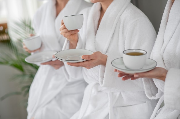 Boire du thé. Mains de femmes en peignoirs blancs avec tasses et soucoupes se reposant dans un spa buvant du thé, visages non visibles