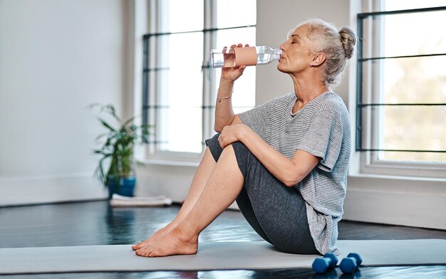 Boire de l'eau fitness et femme âgée sur un tapis de yoga pour l'exercice pilates et l'entraînement à la maison Boire de la relaxation et des haltères pour l'étirement de la mobilité et la pratique de la réadaptation physique pour le bien-être