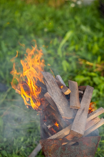 Le bois de chauffage dans le gril brûle avec une flamme de feu orange vif sur un fond vert naturel. Préparation pour la cuisson de la viande sur le gril dans la nature. Flammes de feu et fumée