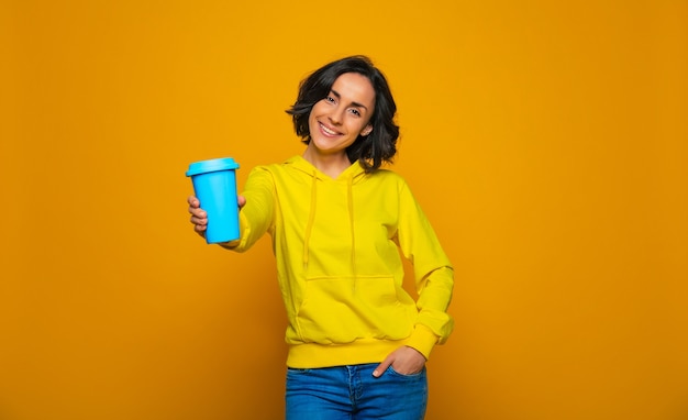 Bois avec moi. smiley fille détendue vêtue d'un sweat à capuche jaune, offrant sa boisson dans une tasse bleue, la tenant sur le bras tendu.