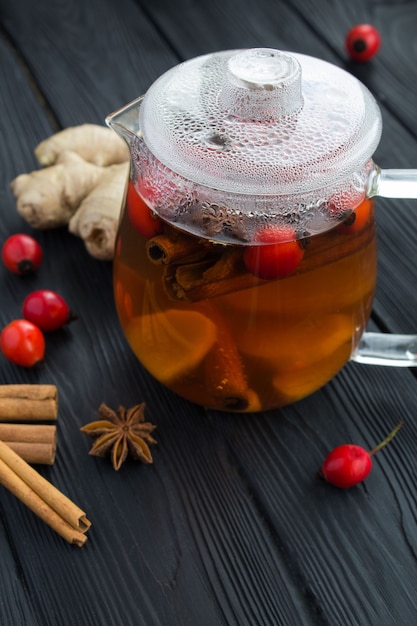 Boisson au thé ou cocktail à l'églantier, au gingembre et aux épices dans la théière en verre sur le fond en bois noir. Emplacement vertical.