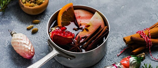 Boisson chaude au vin chaud avec agrumes, pomme, grenade et épices dans une casserole en aluminium avec des jouets d'arbre de noël vintage et une branche de sapin sur fond de béton. Mise au point sélective.