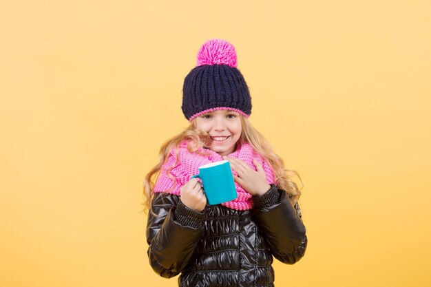 Boisson chaude par temps froid. Enfant avec un sourire de tasse bleue sur fond orange. Concept de détente de la saison d'automne. Fille au chapeau, écharpe rose, veste noire avec mug. Pause thé ou café, espace de copie