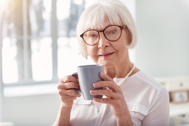 Boisson favorite. Le gros plan d'une charmante femme âgée posant et souriant tout en buvant du café dans une tasse bleue