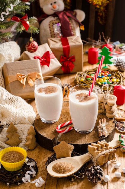 Boisson de lait de poule de Noël et du nouvel an dans des tasses en verre sur un support en bois sur la table avec des cadeaux, des biscuits de Noël, des bougies, des bonbons et des branches d'arbres de Noël.