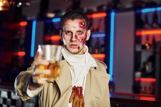 Avec boisson à la main. Portrait d'un homme qui participe à la soirée thématique d'halloween en maquillage et costume de zombie.