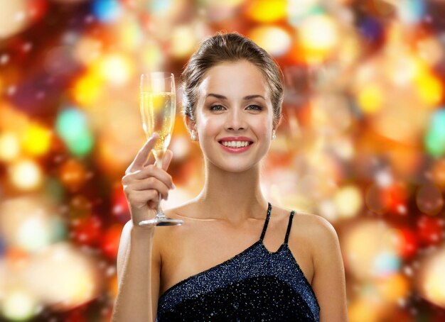 boissons, vacances, noël, personnes et concept de célébration - femme souriante en robe de soirée avec un verre de vin mousseux sur fond rouge de lumières de noël