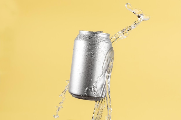 Boîte de bière ou de soda en aluminium avec des éclaboussures d'eau sur un fond jaune clair