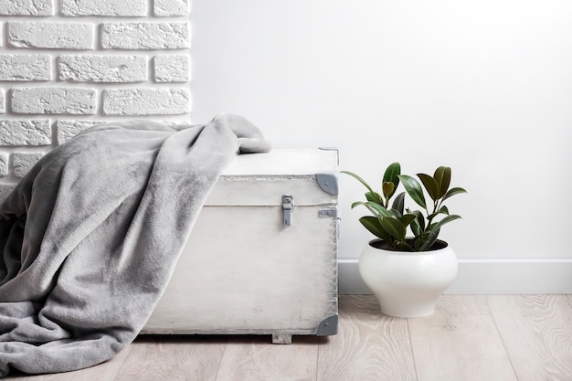 Boîte en bois blanche avec couverture polaire douce grise dessus et jeune plante en caoutchouc dans un pot de fleur blanc. Mur blanc avec des briques sur fond. Copier l'espace