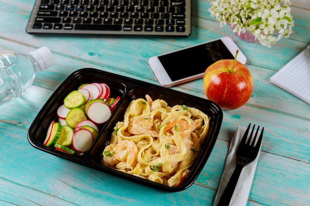 Boîte à lunch contenant des pâtes et des crevettes, du concombre et des radis sur une table en bois avec ordinateur portable.