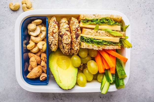 Boîte à lunch saine avec sandwich, biscuits, fruits et avocat sur fond blanc.