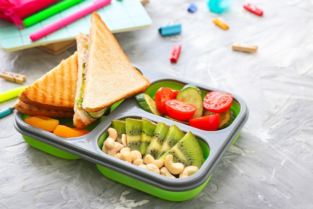 Boîte à lunch scolaire avec de la nourriture savoureuse et de la papeterie sur fond gris