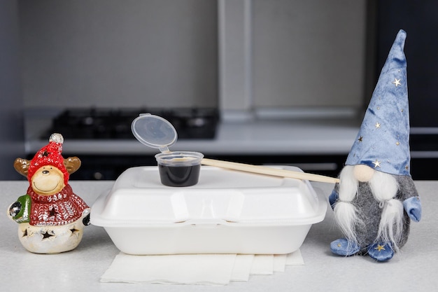 Une boîte sur une table lumineuse à la maison dans la cuisine Sushi de livraison rapide dans un récipient blanc Décoration de figurine Pot de sauce soja