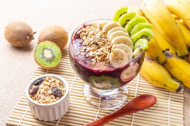 Photo bol d'açai avec kiwi, banane et flocons d'avoine sur une table au décor tropical.