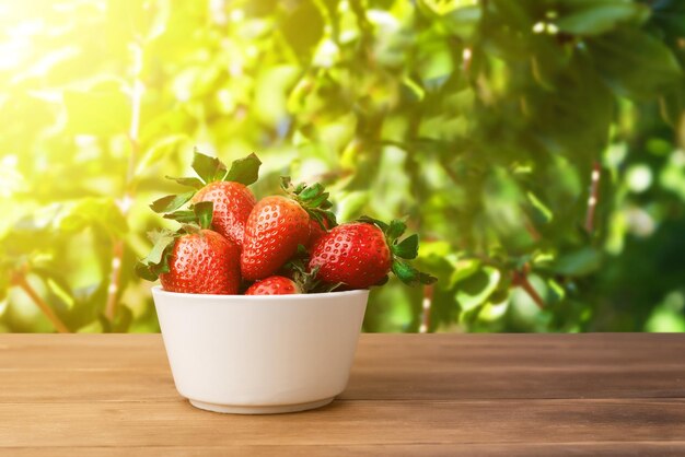 Bol blanc de fraises fraîches sur une table en bois à l'extérieur sur fond de feuilles vertes