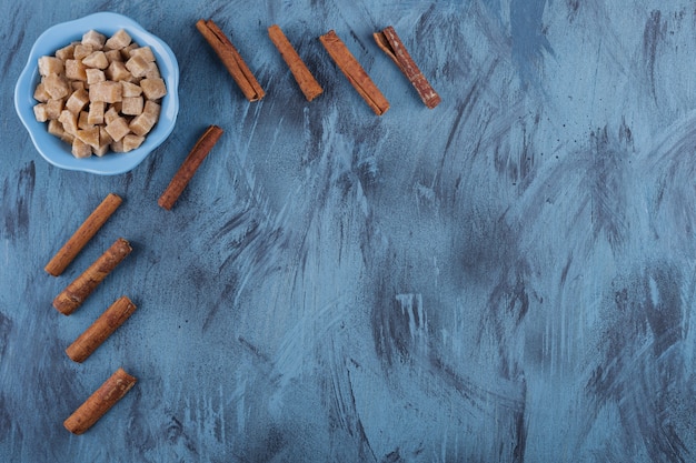 Bol bleu de cubes de sucre brun et de bâtons de cannelle sur une surface bleue.