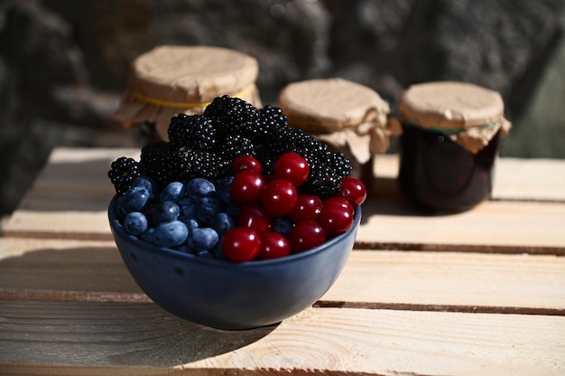 Bol en céramique avec cerises mûres, bleuets et mûres sur une boîte en bois sur fond de confitures assorties floues