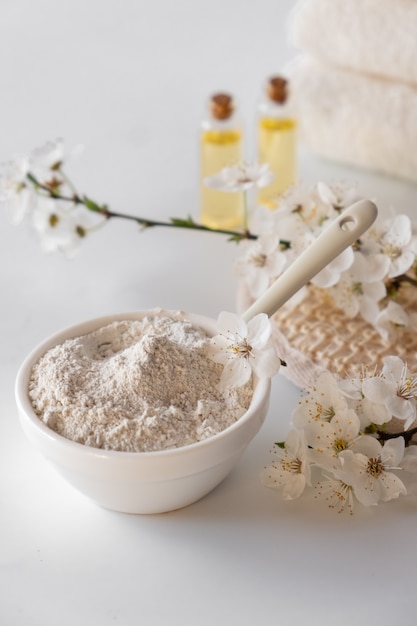 Photo bol en céramique avec de la poudre d'argile blanche sur une surface blanche. ingrédients pour masque ou gommage pour le visage et le corps faits maison et brin frais de cerise en fleurs. concept de spa et de soins corporels.