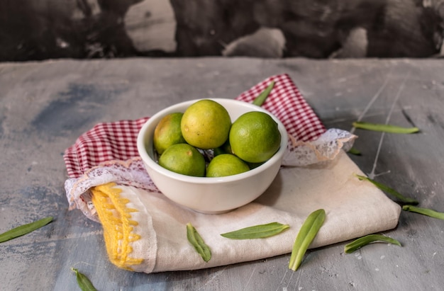 Un bol de citrons verts est posé sur un tissu à carreaux rouges et blancs avec des feuilles vertes dessus.