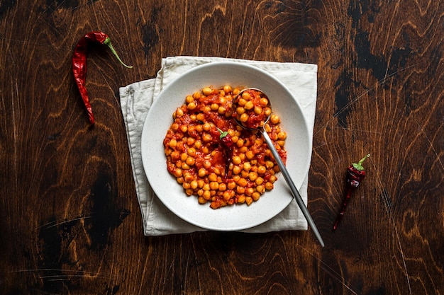 Un bol de compote de pois chiches avec de la sauce tomate chaude sur une serviette, sur fond de bois, vue aérienne. Une cuillère et du piment rouge.