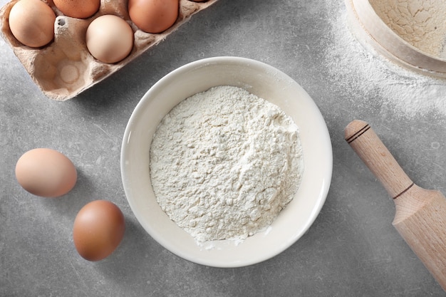 Bol avec farine blanche, œufs et rouleau à pâtisserie sur table de cuisine