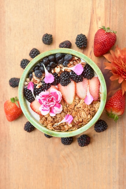 Bol de flocons d'avoine avec granola fraise myrtille et décoré de fleurs sur une table en bois