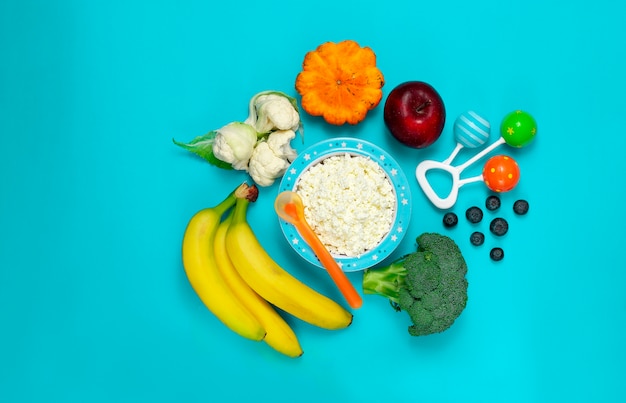 Bol avec fromage cottage, légumes et fruits, nourriture pour bébé, hochet et cuillère, sur fond bleu, vue de dessus, horizontal.