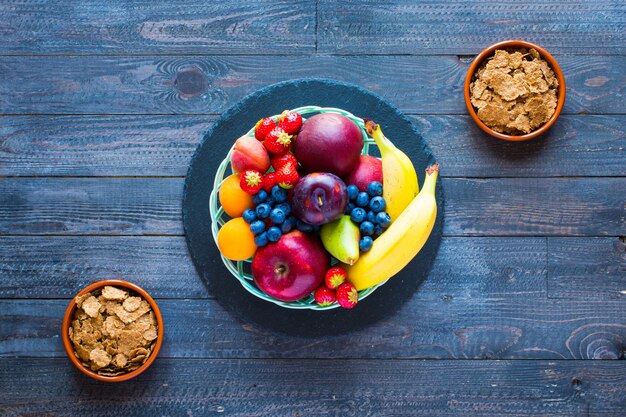 Bol de fruits frais avec banane, pomme, fraises, abricots, myrtilles, prunes, grains entiers, fourchettes, vue de dessus